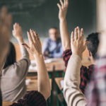 All students have hands raised in classroom for face-to-face teaching