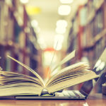 Books and computer are opened on desk in front of library bookshelves