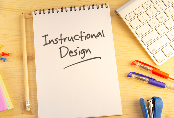 Notebook has the words "Instructional Design Basics" written on it with colored pens and pencils surrounding it