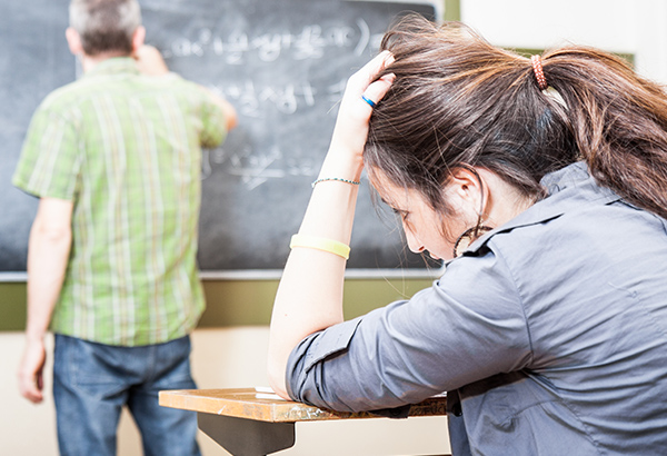 Instructor writes on chalkboard in background while student rests hand on head in desk looking confused