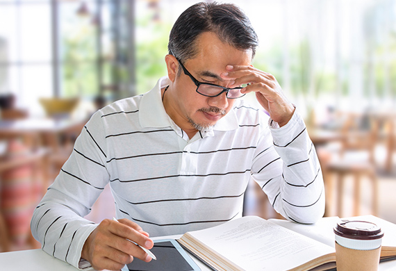 Middle-aged gentleman sits at desk with books and coffee with hand on head for Assimilation Strategies