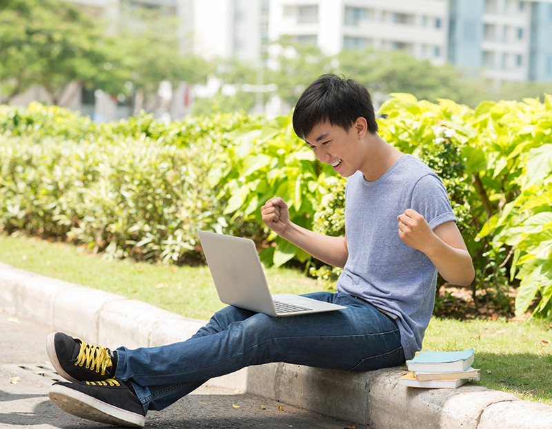 Student celebrates while looking at computer screen sitting outside on campus