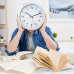 Person holds up clock in replace of their head with books and computer laid out in front of them