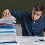 man-in-classroom-at-desk-with-stack-of-papers-folders-building-higher