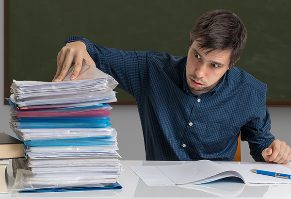 man-in-classroom-at-desk-with-stack-of-papers-folders-building-higher