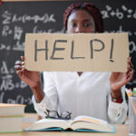 woman-in-classroom-at-desk-holding-up-help-sign