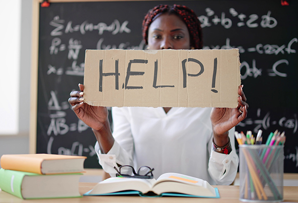 woman-in-classroom-at-desk-holding-up-help-sign