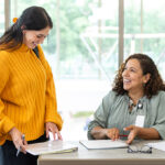 woman-with-backpack-standing-next-to-seated-woman-smiling-and-discussing-papers-in-front-of-them-on-desk