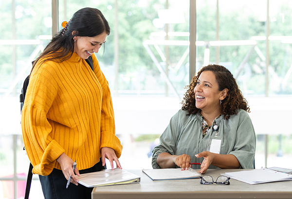 woman-with-backpack-standing-next-to-seated-woman-smiling-and-discussing-papers-in-front-of-them-on-desk